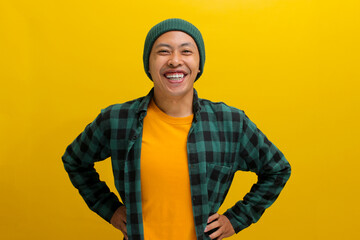 Confident young Asian man stands with a wide smile, his arms akimbo, exuding confidence as he faces the camera against a yellow background