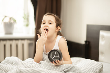 Cute little kid girl sitting on bed waking up yawning in morning and holding alarm clock.