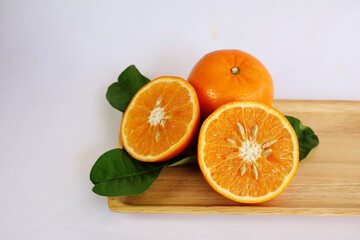 Orange cut in half, flesh and seeds visible, placed on green leaves and wooden tray, white background.