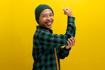 Excited Asian man, dressed in a beanie hat and casual shirt, is making a strong gesture by lifting...
