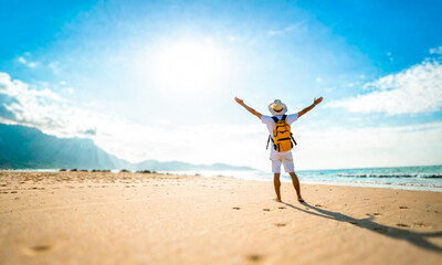 Joyful man, arms wide open, standing on sunny beach, facing ocean. Wearing shorts, hat, carrying backpack. Embracing sea breeze, casual attire, epitomizing leisure, exploration