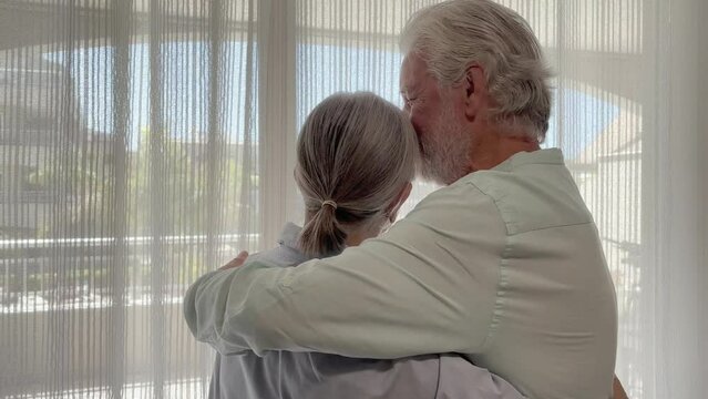 Serene elderly couple at home embracing with affection looking out the window, senior married couple take care of each other and smiling.
