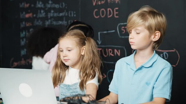 Smart girl using laptop programing engineering code with diverse friends ay table with microchip and main board scatter around in STEM classroom at blackboard written with coding prompt. Erudition.