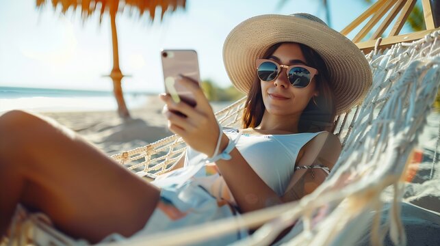 Vacation concept Enjoying the summer Young pretty woman in hat and sunglasses using smartphone laying in hammock on the beach : Generative AI