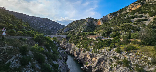 Beautiful view of a deep, rocky mountain gorge in France, with a river, trees, bridge and blue sky.