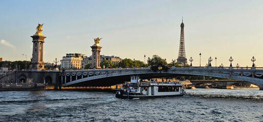 Distant view of the Eiffel Tower, Paris with the River Seine in the foreground, with river cruise boat sailing under a bridge; sunset view of the Eiffel Tower in Paris, France with Pont Alexandre III.