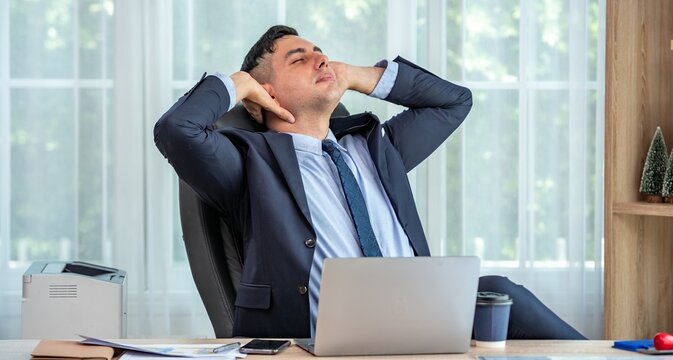 A business man sitting in his office chair with his hands behind his head, leaning back and relaxing.
