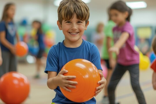 Cheerful young boy holds an orange dodgeball in a gym, with blurred classmates in the background