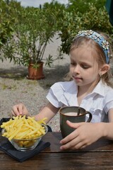 a girl in a headband sits at the table and eats French fries and drinks cocoa from a mug