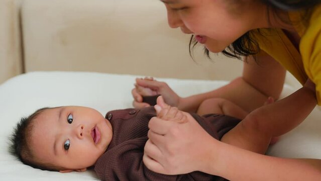 Chubby baby of asian mother playing with newborn smile on bed. mom looking and kiss hands at infant. Holding hand. Talk, touch, Enhance child's development. Happy Family Children, Motherhood.