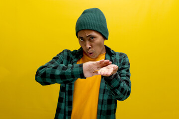 A confident young Asian man, dressed in a beanie hat and a casual shirt, is making a SPEND MONEY gesture while standing against a yellow background