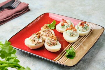 Cold appetizer, eggs stuffed with cream cheese, sun-dried tomatoes, walnuts and herbs on a red ceramic plate on a gray concrete background. Recipes for stuffed eggs.