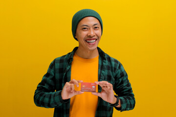 A happy young Asian man, dressed in a beanie hat and casual shirt, enthusiastically shows a credit...