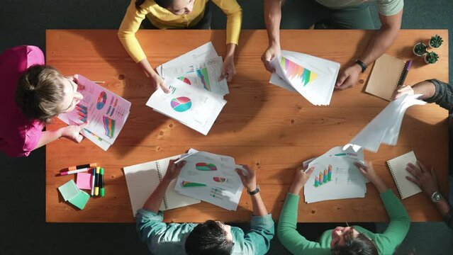 Top view of smart people throw paper and giving high five at meeting table with financial document and stock market analysis paper scattering around. Aerial view of team celebrate project. Symposium.