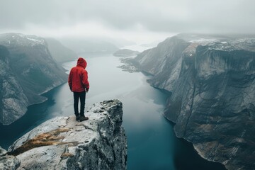 A man in a red jacket stands on a rocky ledge overlooking a body of water - Powered by Adobe