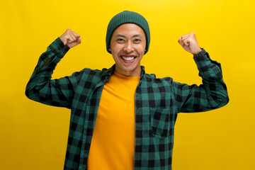 Excited Asian man, dressed in a beanie hat and casual shirt, is making a strong gesture by lifting his arm to show muscles, expressing pride in his achievements, standing against a yellow background