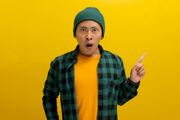 Astonished Asian man, dressed in a beanie hat and casual shirt, points at an empty space, possibly...