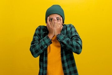 Fearful young Asian man, dressed in a beanie hat and casual shirt, covers his eyes, possibly shielding himself from the frightful or overwhelming scenes while watching horror movies during Halloween