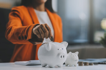 Beautiful Asian businesswoman insert coins in a pin piggy bank on the table. saving money,...