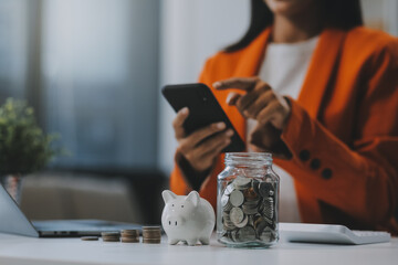 Beautiful Asian businesswoman insert coins in a pin piggy bank on the table. saving money,...