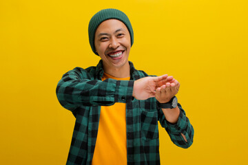 A confident young Asian man, dressed in a beanie hat and a casual shirt, is making a SPEND MONEY gesture while standing against a yellow background