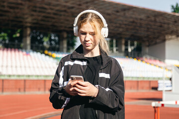 Teenager girl uses a smartphone and listens to music on headphones at the stadium, outdoor workout...