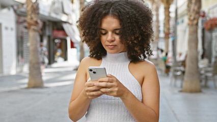 A young hispanic woman with curly hair using a smartphone on a sunny city street