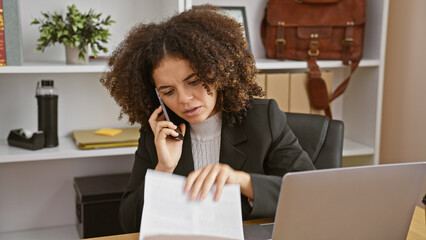 A young hispanic woman with curly hair, engaged in a phone conversation while working in an indoor...