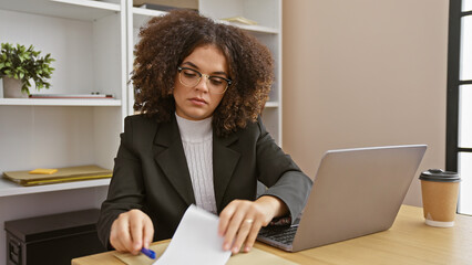 A young hispanic woman with curly hair, wearing glasses, works on her laptop in a modern office...