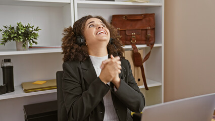 A hopeful young hispanic woman with curly hair prays in an office setting, embodying optimism.