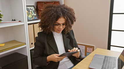 A young hispanic woman with curly hair using a tablet in a modern office setting.