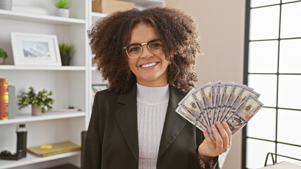 Happy young hispanic woman with curly hair holding us dollars in modern office, showcasing success...