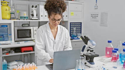 Hispanic woman scientist working on laptop in laboratory indoors surrounded by research equipment.