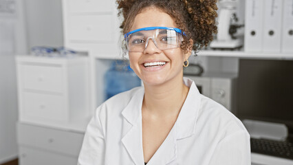 A smiling young hispanic woman with curly hair wearing safety glasses and a lab coat in a clinic.