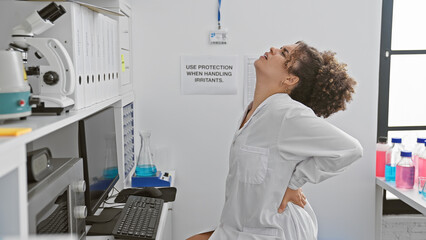 A young woman with curly hair wearing a white lab coat stretches her back in an indoor laboratory...