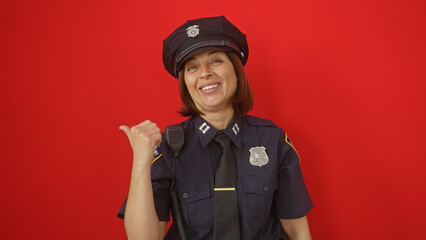 Smiling middle-aged hispanic female police officer gesturing with thumb against a red background.