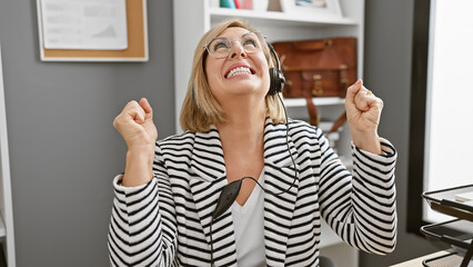 A joyful middle-aged blonde woman in an office, wearing a headset, celebrates success with a...