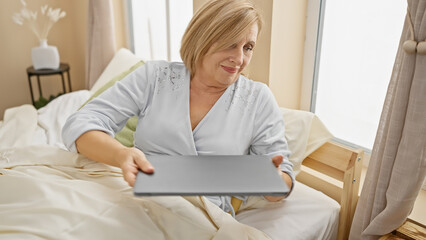 Mature caucasian woman in bedroom holding laptop with a contemplative expression at home