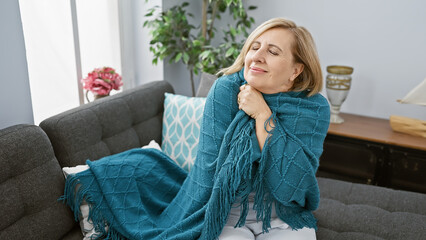 Blonde woman relaxing on a couch at home draped in a teal throw, eyes closed with a contented smile.