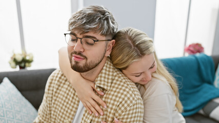 A contented young man and woman embrace tenderly on a couch in a cozy living room, symbolizing love and togetherness.