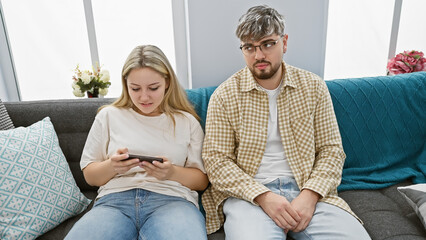 Young couple lounging on sofa with woman focused on smartphone and man looking away thoughtfully in...