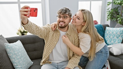 A loving couple takes a selfie indoors on the sofa, showcasing their relationship in a cozy home...