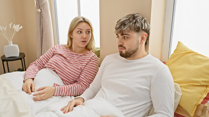A surprised woman and a confused man sitting together on a bed in a well-lit bedroom, portraying a couple potentially having a conversation.