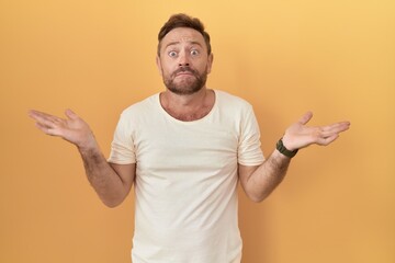 Middle age man with beard standing over yellow background clueless and confused expression with...