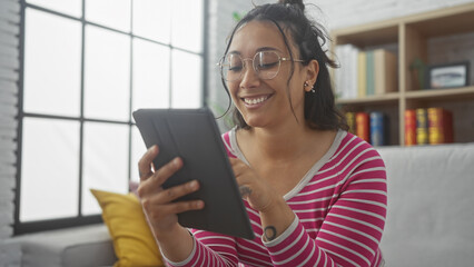 Smiling young hispanic woman using tablet in modern living room with bookshelf background.