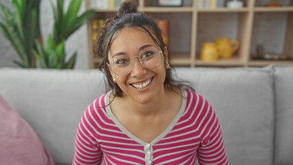 A smiling young hispanic woman with glasses seated at home exuding a casual and approachable vibe.