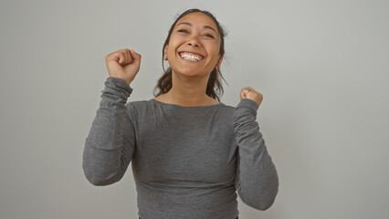 Joyful young hispanic woman celebrating with clenched fists against a plain white wall in a casual...