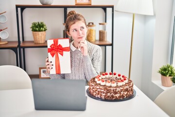 Thoughtful young blonde woman celebrating with a gift, sitting at home expressing serious face, her...