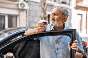 Senior grey-haired man drinking coffee standing by car at street