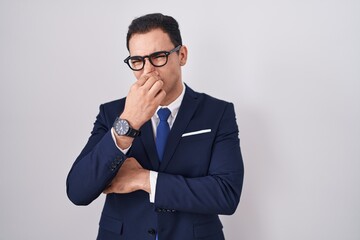 Young hispanic man wearing suit and tie smelling something stinky and disgusting, intolerable smell, holding breath with fingers on nose. bad smell